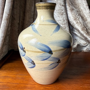 May include: A ceramic vase with a beige base and a flared top. The vase is adorned with blue leaf-like patterns and brown accents. The rim is a darker shade of brown. The vase is sitting on a wooden surface.