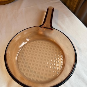 May include: An amber-coloured glass skillet with a handle. The skillet has a round shape and a textured base. The rim of the skillet is black. The handle is an extension of the skillet's body.