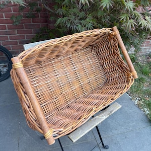 May include: A rectangular wicker basket with a natural brown finish, featuring a woven base and sides. The basket is constructed with thicker, smooth wooden rods at the top corners and is supported by a black metal folding stand with a wooden crossbar.