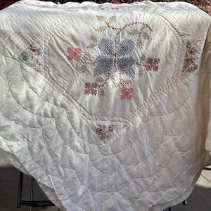 May include: Cream-coloured, quilted blanket with an embroidered butterfly design in blue, pink, and green. The blanket has a scalloped edge and floral patchwork accents, showcasing a vintage aesthetic.