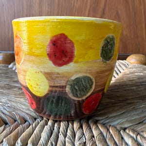 May include: A hand-painted ceramic bowl with a yellow background and colourful polka dots in red, green, orange, and yellow. The bowl has a brown base and a slightly textured surface. The bowl is sitting on a woven surface.