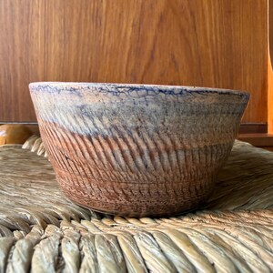 May include: A handcrafted ceramic bowl with a textured, ridged exterior. The bowl has a gradient of colours, shifting from a light blue rim to a brown base. The bowl is on a woven surface.