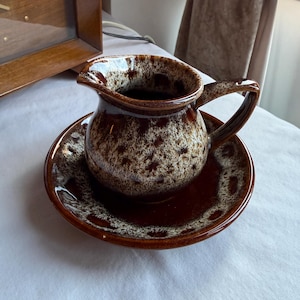 May include: A brown ceramic creamer and saucer set. The creamer has a handle and spout, and the set features a speckled pattern in shades of brown and cream. The creamer sits on a matching saucer.