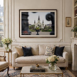 Könnte beinhalten: Ein gerahmtes Foto einer Kathedrale mit Türmen hängt über einem beigen Sofa mit Zierkissen. Der Raum verfügt über einen Kamin, einen gemusterten Teppich und einen goldgerahmten Spiegel. Eine Glasvase enthält weiße Blumen.