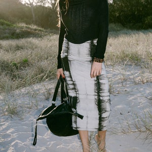 May include: A woman wearing a black mock neck sweater and a white and black tie-dye skirt. She is holding a black leather tote bag and standing on a sandy beach.