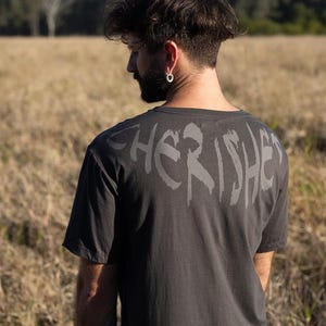May include: Dark grey t-shirt with the word "CHERISHED" printed across the back in a stylised font. The shirt is worn by a person standing in a field of dry grass.