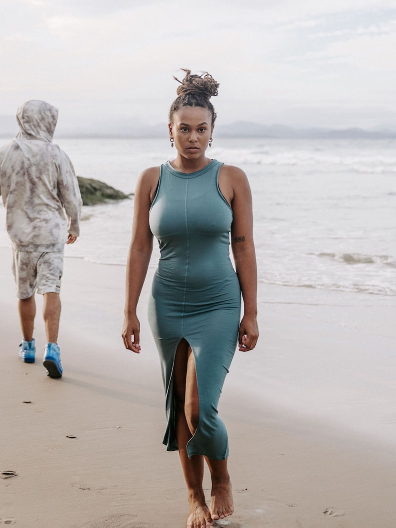 Peut inclure: Une femme portant une robe maxi sans manches bleu sarcelle avec une fente sur le devant, marchant sur une plage de sable.