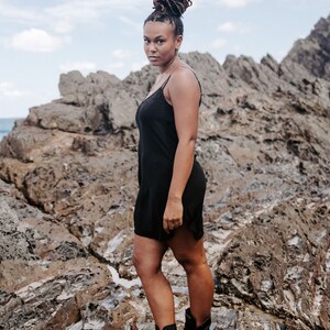May include: A woman wearing a black slip dress and black combat boots stands on a rocky surface. She has dark brown hair styled in a bun.