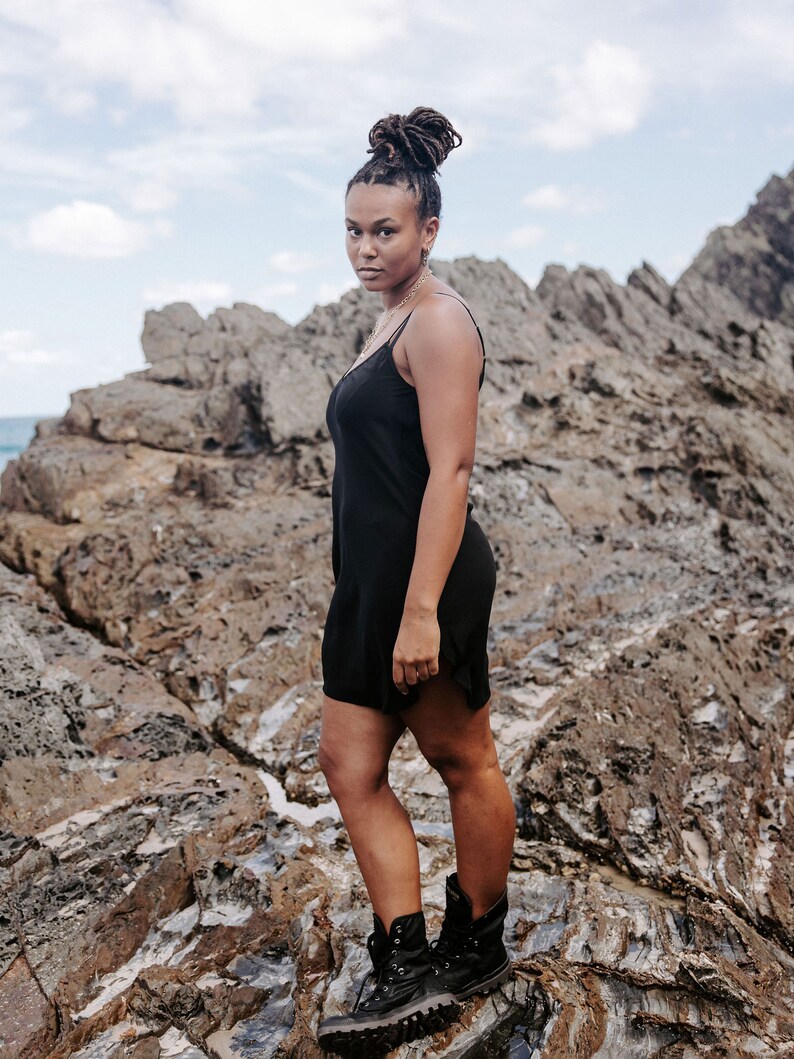 May include: A woman wearing a black slip dress and black combat boots stands on a rocky surface. She has dark brown hair styled in a bun.