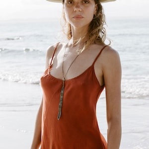 May include: A woman wearing a rust-colored slip dress with a long, thin pendant necklace. She is standing on a beach and wearing a green hat.