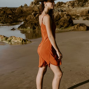 May include: A woman wearing a rust-colored slip dress and brown leather boots stands on a sandy beach. She is wearing a green felt fedora hat with a brown leather band.