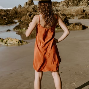 May include: A woman wearing a rust-colored slip dress and a brown fedora stands on a sandy beach. She is looking out at the ocean.