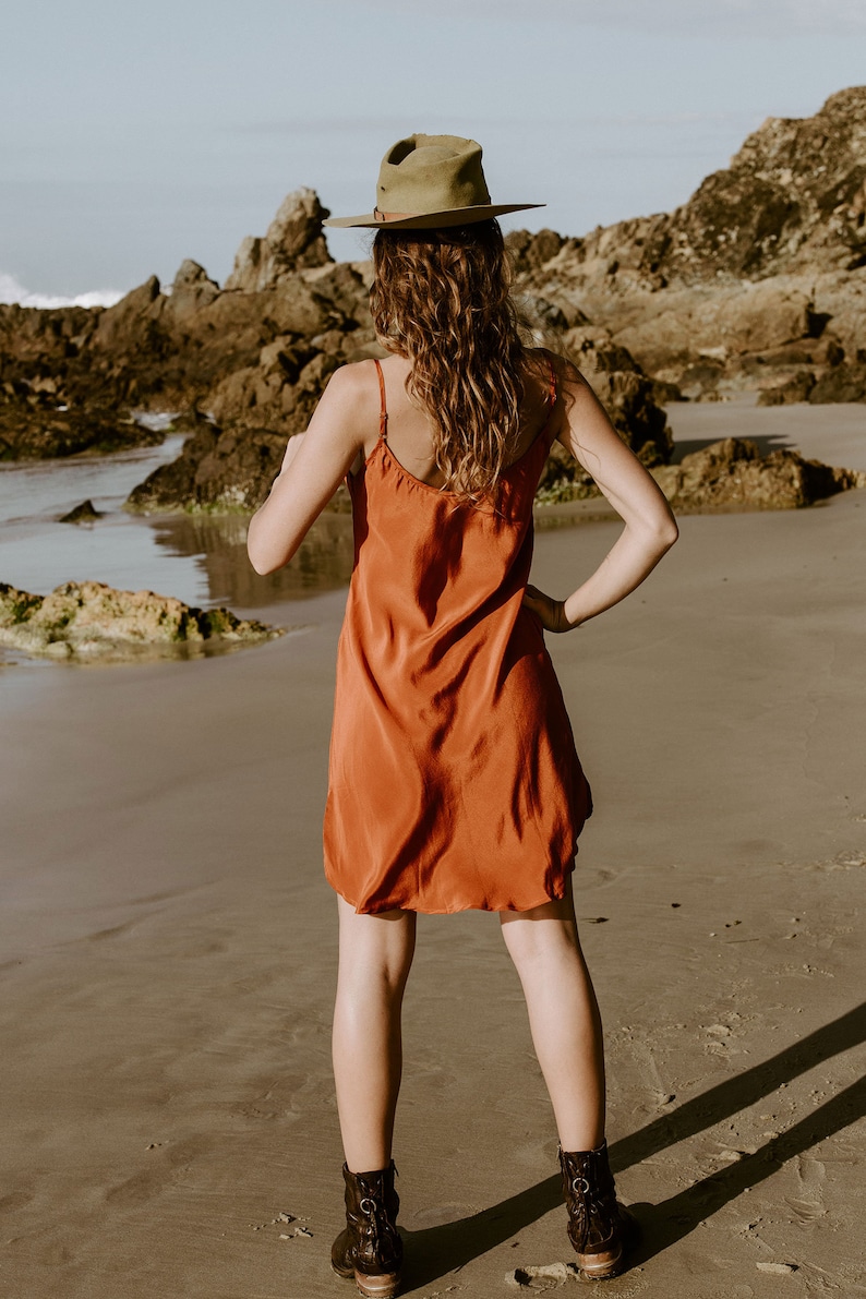 May include: A woman wearing a rust-colored slip dress and a brown fedora stands on a sandy beach. She is looking out at the ocean.