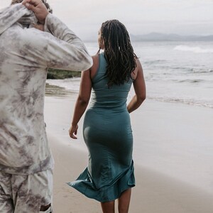 Peut inclure: Une personne portant une chemise &agrave; motifs blancs et gris marche sur une plage de sable. Une personne portant une robe maxi turquoise marche devant elle, regardant l'oc&eacute;an.