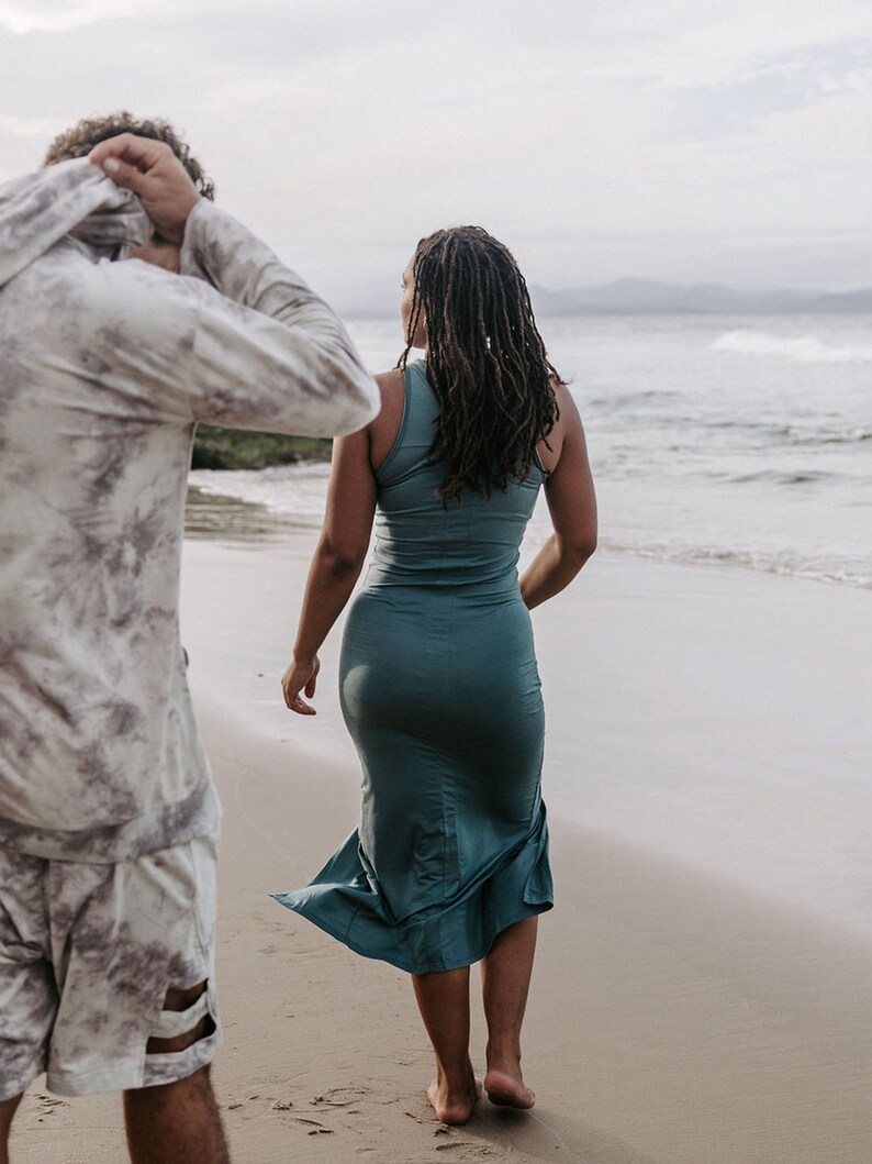 Peut inclure: Une personne portant une chemise &agrave; motifs blancs et gris marche sur une plage de sable. Une personne portant une robe maxi turquoise marche devant elle, regardant l'oc&eacute;an.