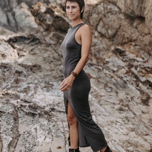 Op de afbeelding: Een vrouw in een donkergrijze maxi-jurk met een zijsplit en bruine leren enkellaarsjes. Ze staat op een rotsachtig strand.