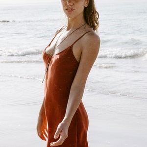 May include: A woman wearing a rust-colored slip dress and a light green fedora hat stands on a sandy beach. The ocean is in the background.