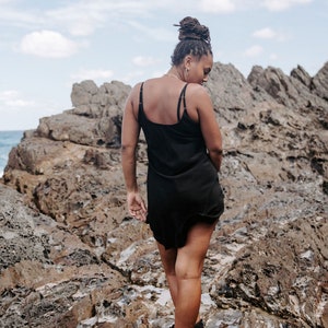 May include: A person wearing a black slip dress and black boots stands on a rocky surface. The person has their back to the camera and is looking down.