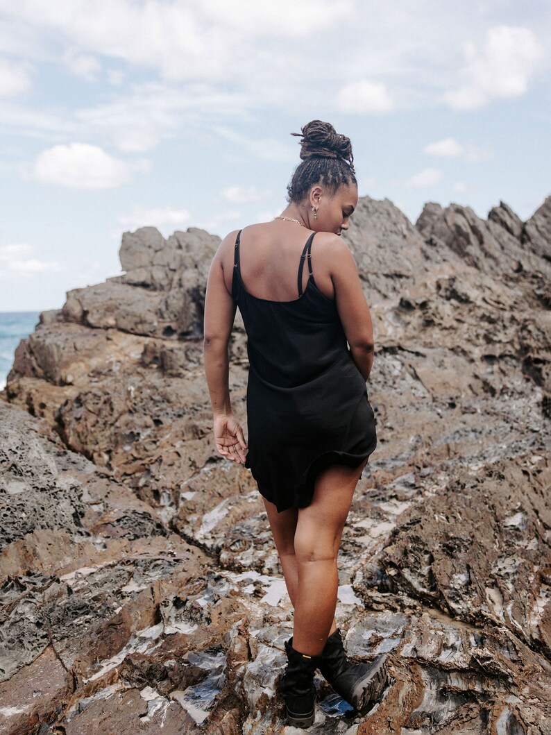 May include: A person wearing a black slip dress and black boots stands on a rocky surface. The person has their back to the camera and is looking down.