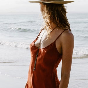May include: A woman wearing a rust-colored slip dress with a long, silver pendant necklace. She is standing on a sandy beach with the ocean in the background. She is wearing a light brown fedora.