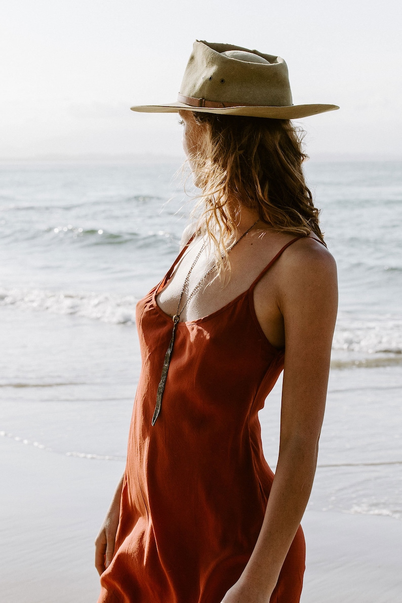 May include: A woman wearing a rust-colored slip dress with a long, silver pendant necklace. She is standing on a sandy beach with the ocean in the background. She is wearing a light brown fedora.