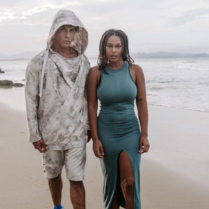 Peut inclure: Un couple debout sur une plage de sable. L'homme porte un sweat &agrave; capuche avec un motif tie-dye et des baskets bleues. La femme porte une robe sans manches turquoise avec une fente sur le c&ocirc;t&eacute;.