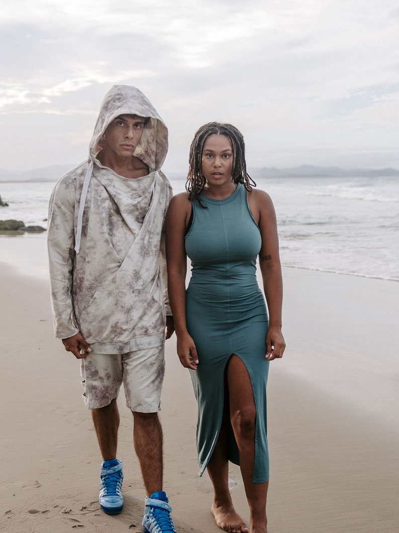 Peut inclure: Un couple debout sur une plage de sable. L'homme porte un sweat &agrave; capuche avec un motif tie-dye et des baskets bleues. La femme porte une robe sans manches turquoise avec une fente sur le c&ocirc;t&eacute;.