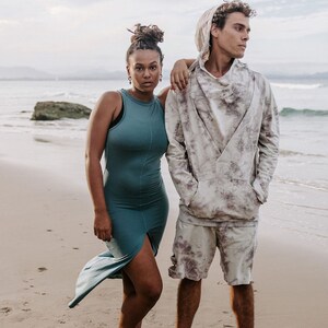 Peut inclure: Un couple debout sur une plage de sable. La femme porte une robe sans manches turquoise et l'homme porte un sweat &agrave; capuche blanc et gris tie-dye et un short assorti. Ils portent tous les deux des baskets bleues.