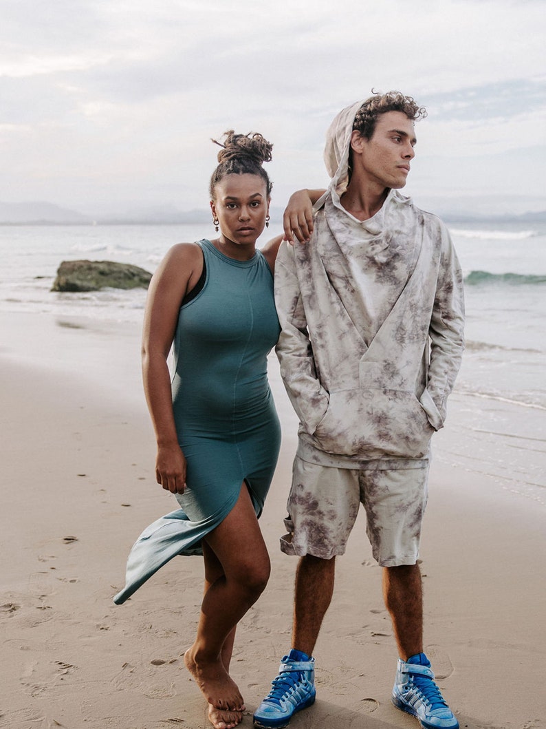 Peut inclure: Un couple debout sur une plage de sable. La femme porte une robe sans manches turquoise et l'homme porte un sweat &agrave; capuche blanc et gris tie-dye et un short assorti. Ils portent tous les deux des baskets bleues.