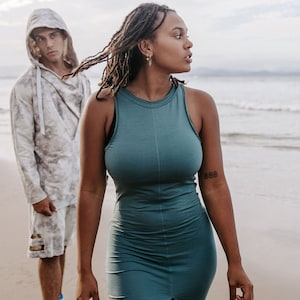 Peut inclure: Une femme avec de longues dreadlocks porte une robe sans manches bleu sarcelle avec une fente sur le devant. Elle se tient sur une plage avec un homme portant un sweat &agrave; capuche blanc et un short derri&egrave;re elle.