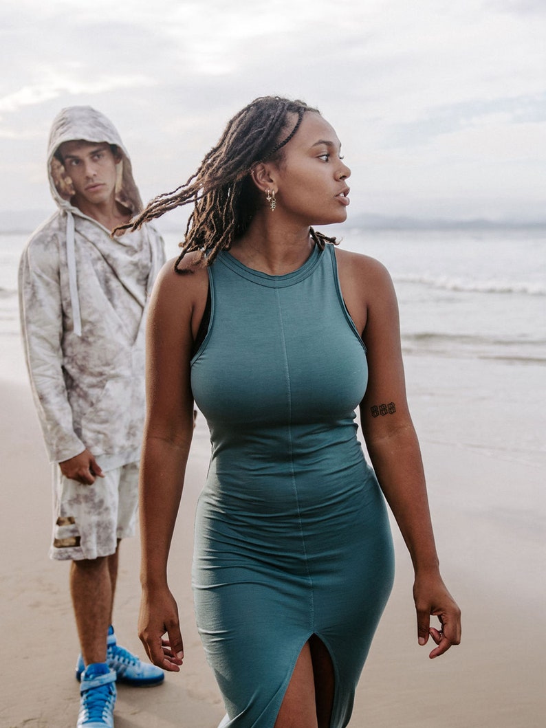 Peut inclure: Une femme avec de longues dreadlocks porte une robe sans manches bleu sarcelle avec une fente sur le devant. Elle se tient sur une plage avec un homme portant un sweat &agrave; capuche blanc et un short derri&egrave;re elle.