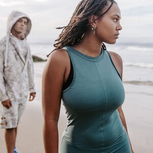 Peut inclure: Une femme avec de longues dreadlocks portant une robe sans manches turquoise se tient sur une plage, regardant de c&ocirc;t&eacute;. Un homme portant un sweat &agrave; capuche et un short se tient derri&egrave;re elle.