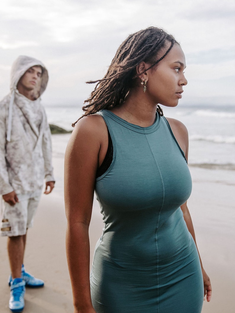 Peut inclure: Une femme avec de longues dreadlocks portant une robe sans manches turquoise se tient sur une plage, regardant de c&ocirc;t&eacute;. Un homme portant un sweat &agrave; capuche et un short se tient derri&egrave;re elle.
