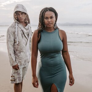 Peut inclure: Un homme portant un sweat &agrave; capuche et un short se tient &agrave; c&ocirc;t&eacute; d'une femme portant une robe sans manches turquoise. Ils sont tous les deux sur une plage de sable avec l'oc&eacute;an en arri&egrave;re-plan.