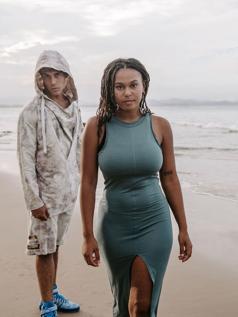 Peut inclure: Un homme portant un sweat &agrave; capuche et un short se tient &agrave; c&ocirc;t&eacute; d'une femme portant une robe sans manches turquoise. Ils sont tous les deux sur une plage de sable avec l'oc&eacute;an en arri&egrave;re-plan.