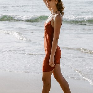 May include: A woman wearing a rust-colored slip dress and a brown hat walks on a sandy beach. She is wearing brown ankle boots.