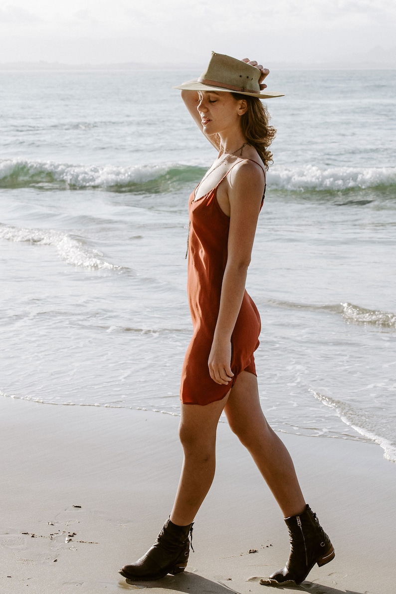 May include: A woman wearing a rust-colored slip dress and a brown hat walks on a sandy beach. She is wearing brown ankle boots.