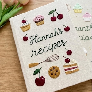Könnte beinhalten: Ein cremefarbenes Rezeptbuch mit dem gestickten Text "Hannah's recipes". Das Cover zeigt gestickte Cupcakes, Kirschen, einen Schneebesen, einen Keks und ein Stück Kuchen. Das Buch ist geschlossen, mit einem Leseband.