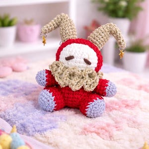 May include: A handmade crochet jester doll in red, white, and tan. The doll has a red body, white face, and tan jester hat with gold star charms. The doll is sitting on a colorful, fluffy blanket.