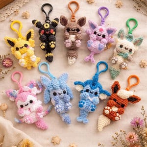 May include: A collection of handmade crochet keychains in various colors, featuring cartoon animal designs. The keychains have metal rings and are arranged on a cream-colored surface with floral accents. Each keychain is approximately 3 inches tall.
