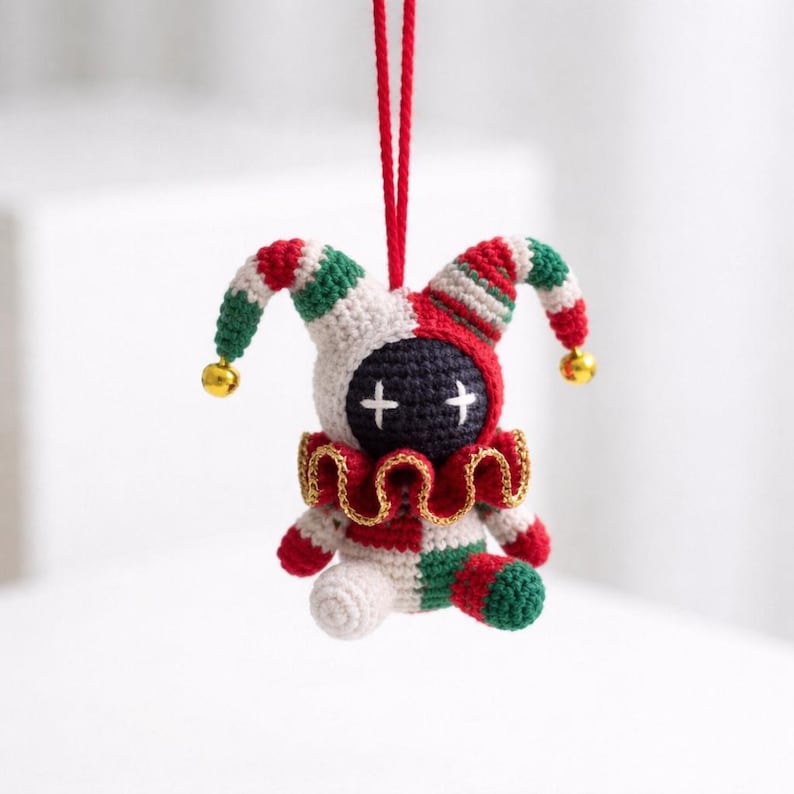 May include: A handmade crochet jester doll ornament. The doll features a black face with white cross eyes, a red, white, and green jester hat, and a ruffled collar. Gold bells hang from the hat. The doll is suspended by a red string.
