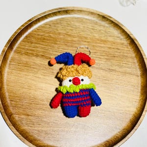 May include: A handmade crochet clown keychain. The clown has a red nose, a white face, and a yellow, blue, and red outfit. The keychain is on a wooden tray with a natural wood grain.