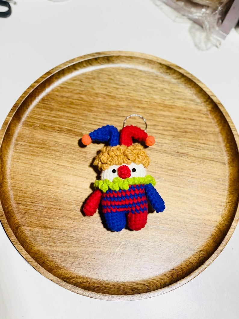May include: A handmade crochet clown keychain. The clown has a red nose, a white face, and a yellow, blue, and red outfit. The keychain is on a wooden tray with a natural wood grain.