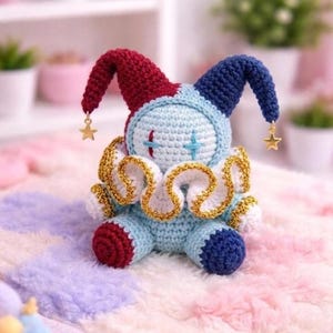 May include: A handmade crochet jester doll in shades of blue, red, and white. The doll has a light blue body, red and blue pointed hat, and a gold-trimmed collar. Gold star charms dangle from the hat. The doll is sitting on a soft, multi-colored surface.
