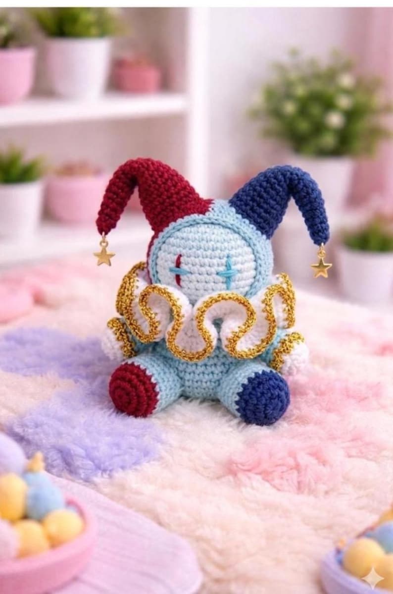 May include: A handmade crochet jester doll in shades of blue, red, and white. The doll has a light blue body, red and blue pointed hat, and a gold-trimmed collar. Gold star charms dangle from the hat. The doll is sitting on a soft, multi-colored surface.