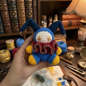 May include: A handmade crochet doll in blue, yellow, red, and white, resembling a jester. The doll has long blue ears with gold star charms, a red ruffled collar, and yellow feet and hands. The doll is held in front of a bookshelf.