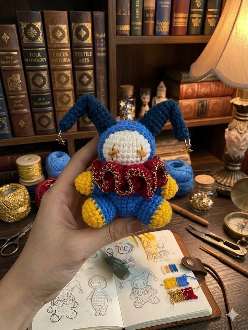 May include: A handmade crochet doll in blue, yellow, red, and white, resembling a jester. The doll has long blue ears with gold star charms, a red ruffled collar, and yellow feet and hands. The doll is held in front of a bookshelf.