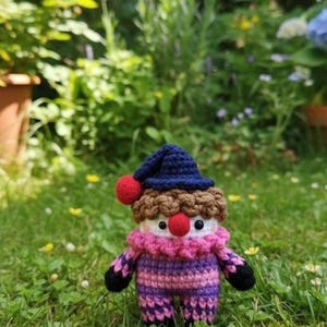 May include: A handmade crochet doll in a garden setting. The doll has a blue hat with a red pom-pom, brown hair, and a pink and purple striped outfit. The doll's face has red nose and black eyes. The doll is standing in green grass.