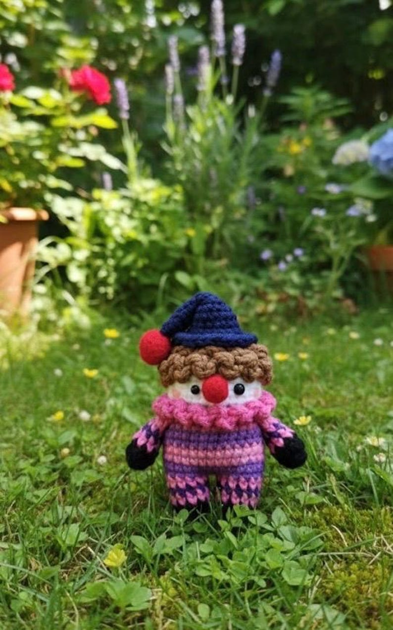 May include: A handmade crochet doll in a garden setting. The doll has a blue hat with a red pom-pom, brown hair, and a pink and purple striped outfit. The doll's face has red nose and black eyes. The doll is standing in green grass.