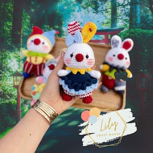May include: Crocheted clown and bunny dolls. The bunny doll is white with a red nose, blue bow, and a yellow and red striped ear. The clown dolls have red noses and colorful outfits. The dolls are handmade and displayed on a wooden tray.